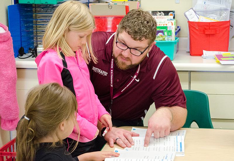 A male teacher helps two female elementary students in a classroom.